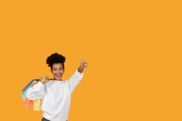 A joyful woman wearing a white sweatshirt holds multiple colorful shopping bags while pointing...