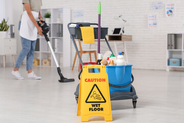 Janitor with trolley, caution sign and vacuum cleaner cleaning floor in office