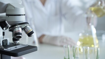 A scientist, wearing a lab coat and white protective gloves, is pouring a yellow oily liquid from one beaker to another near microscope in laboratory, close up. Medicine and science concept