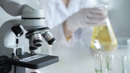 Microscope is near a scientist, wearing a lab coat and white protective gloves, and holding beaker with a yellow oily liquid in laboratory setting, close up. Medicine and science concept