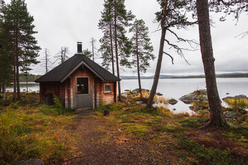 Wooden shelter called kota next to the shore of Inari lake with trees in late autumn wet nature provides safety, relax place and warmth for hikers in harsh arctic condition in Lapland, Finland