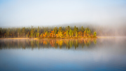 Fog over the finnish lake during cold morning hides the colorful golden trees which reflect in the calm water in Lapland evokes calm, cold, start of the day, solitude, peaceful, Finland