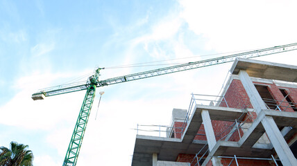 Construction crane tower rises above skyline with workers maneuvering equipment at a busy urban site during daylight hours