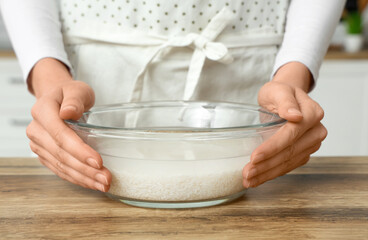 Woman holding glass bowl of water and rice near table in kitchen, closeup