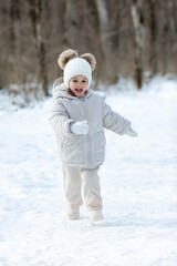 Happy little girl in warm white jacket and white knitted hat has fun and walking in snowy winter park or forest. Children outdoor activities concept. Happy family time