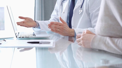 Doctor is consulting patient in modern fair clinic. Healthcare professional discusses medical information with a woman, gesturing above the laptop computer on the glass desk. Medicine