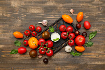 Board with different fresh tomatoes, basil, garlic and bowl of salt on wooden background
