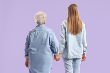 Senior woman and her granddaughter holding hands on purple background, back view