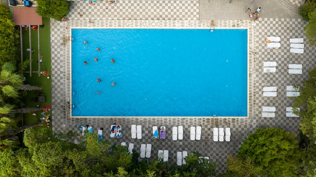 Aerial perpendicular view of a rectangular pool with people bathing. Around the pool there are other people and sun loungers on a beautiful decorated floor.
