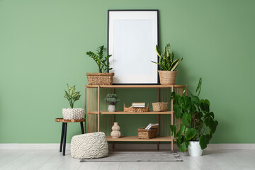 Shelving unit, houseplants, pouf and frame near green wall in room