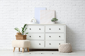 Chest of drawers, poufs, houseplants and frames near white brick wall in room
