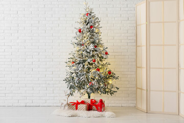 Decorated Christmas tree with glowing lights and gift boxes near white brick wall