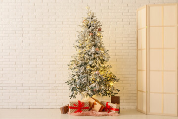 Decorated Christmas tree with glowing lights and gift boxes near white brick wall