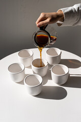 Pouring freshly brewed coffee into simple white cups on a minimalist table illuminated by soft natural light
