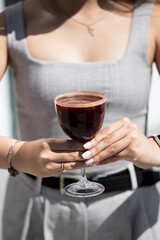 A woman holding a glass of dark drink outdoors on a sunny day, showcasing her stylish outfit and well-manicured nails