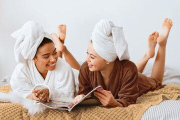 Young beautiful multiracial women in bathrobes and towels on head happily talking and reading...