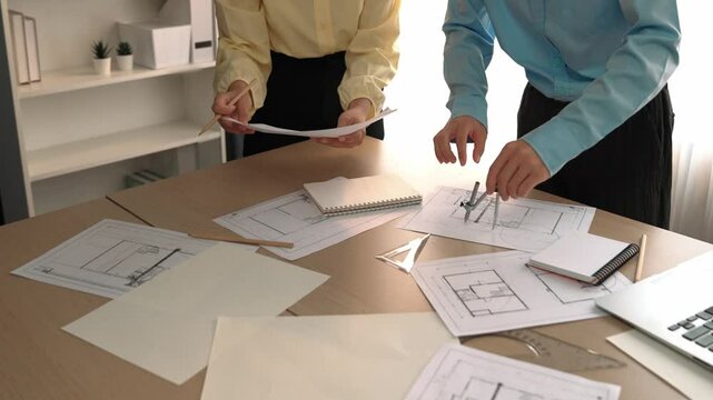 Architect and safety engineer discussion about building blueprint displaying on laptop on table with architectural document and wooden block scatter around at modern office. Closeup. Delineation.
