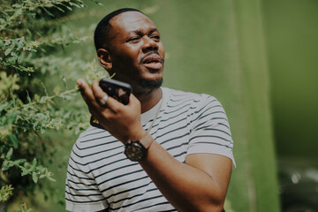 A man stands outside under the sun, engaging with his smart phone amidst greenery. He appears thoughtful and attentive, enjoying the natural ambiance while focusing on his device.