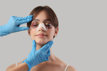 Surgeon hands touching happy woman's bandaged nose after plastic surgery on grey background