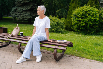 Full length portrait of senior woman finding peace and tranquility in lush park, seated on bench with coffee and backpack, embracing contentment and relaxation of beautiful summer day
