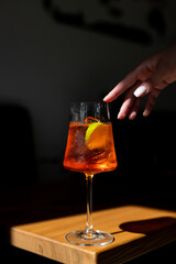 A hand reaches for a refreshing orange cocktail garnished with citrus in a dimly lit setting during the early evening