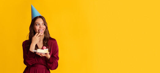 Pensive young lady in party hat holding birthday cake, eating cream, licking finger with pleasure and looking aside at copy space on colorful background