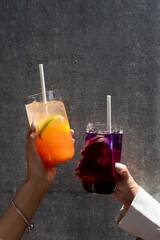 Two hands raising colorful drinks against a textured wall in a casual setting during daylight