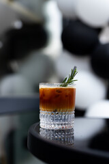 A stylish glass of iced coffee with rosemary garnish placed on a sleek black table against a backdrop of elegant balloons