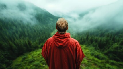 A lone hiker in a red jacket gazes at a breathtaking foggy mountain vista, standing atop a green slope, reflecting on solitude and the beauty of nature.