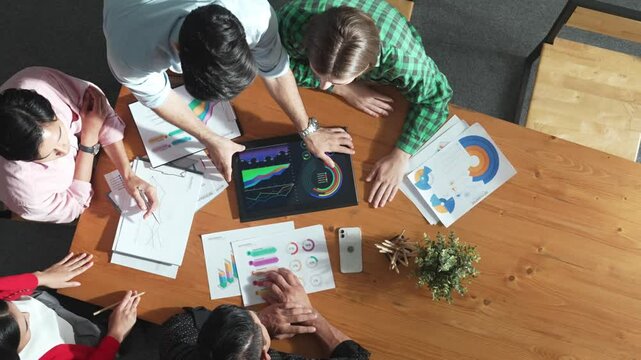 Top view of business man using tablet display increasing graph while diverse marketing people sharing idea or planing strategy at meeting table with stock market statistic scatter around. Convocation.