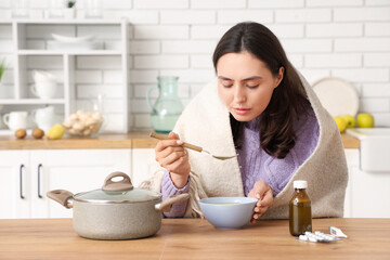 Sick young woman eating soup at table in kitchen