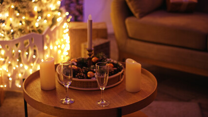 Two champagne glasses stand on a coffee table by a decorated christmas tree with burning candles, creating a cozy and festive atmosphere