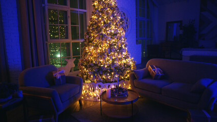 Large christmas tree is standing in a living room, illuminating presents placed on the sofas next to it. The room is decorated for the holidays