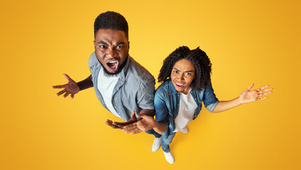 Top View Of Angry Black Couple Emotionally Reacting To Something, Objecting With Raised Hands, Standing Together On Yellow Studio Background