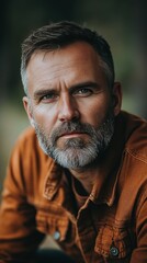 Fototapeta premium A mature man with a well-groomed beard gazes thoughtfully into the distance. He wears a brown jacket and is surrounded by soft natural light that enhances the moment