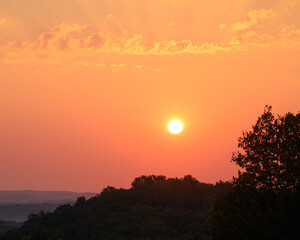 A brilliant orange sun rising over the forested hills of southern indiana. Beautiful and serene scene. Simple composition with sun, clouds, and forest