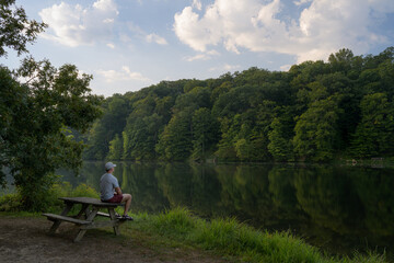 Man sitting on a picnic table overlooking a lake surrounded by a forest. Early morning light with a blue sky with puffy clouds. Summer serene and tranquil moment