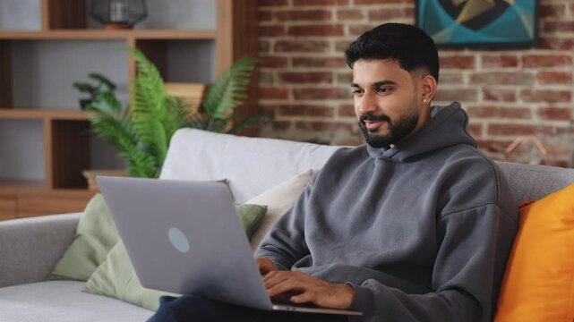Overjoyed dark haired man holding computer on laps and cheering with victory gesture in renovated apartment. Excited male receiving email with food news and celebrating receiving approval.