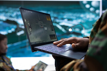 Intelligence specialist monitoring a world map and surveillance imagery on laptop, using the satellite radar system with cctv. Soldiers manage reconnaissance activity in army command post.