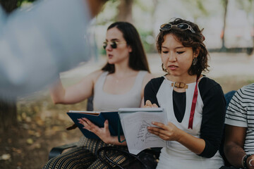 A group of young business people is engaged in a collaborative meeting outdoors. They are discussing ideas, taking notes, and enjoying a productive work session in a relaxed environment.