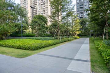 Modern Residential Complex Garden with Wide Paved Walkway and Manicured Landscapes with Mixed Vegetation, Including Ground Cover Plants and Tree-Lined Pathways in communal outdoor space of apartments