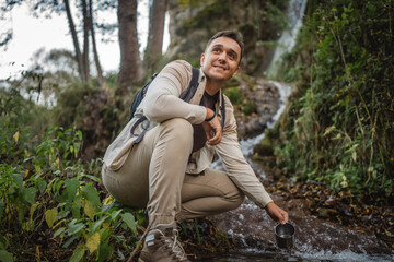 Adult man hiker backpacker squats by a river, filling a mug with water