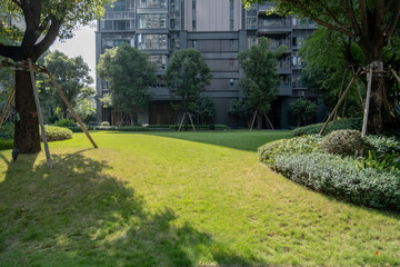 A well-manicured green lawn or grassy area surrounded by landscaped trees and shrubs.A modern apartment building in the background. An outdoor recreational or common area within a residential complex.
