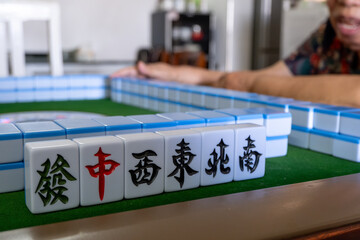 A row of six mahjong tiles in the center reads 'get rich', 'center 'West,' 'East,' 'North,' and 'South' in Chinese characters, against a blurry background of an elderly woman sitting behind the table.