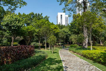 A winding paved pathway meanders through a lush, green urban park with trimmed hedges, shrubs and trees. Some modern high-rise apartment buildings in the background.