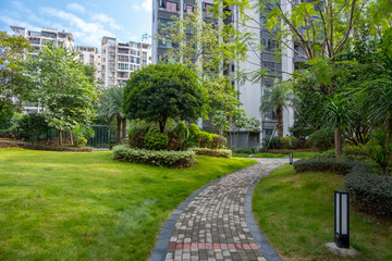 A serene courtyard garden or communal green outdoor space in a modern residential apartment building complex, featuring a well-manicured lawn, lush greenery, and a winding brick pathway.