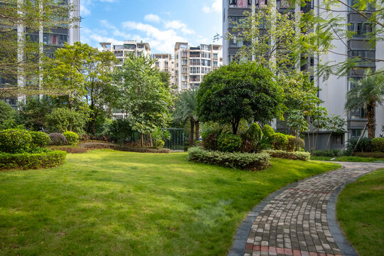 A serene courtyard garden or communal green outdoor space in a modern residential apartment building complex, featuring a well-manicured lawn, lush greenery, and a winding brick pathway.