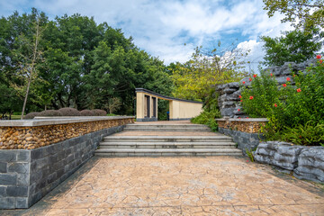 A stone-paved walkway leads up to a set of steps with decorative landscaping, including stone retaining walls, plants, and flowering bushes in a public park.