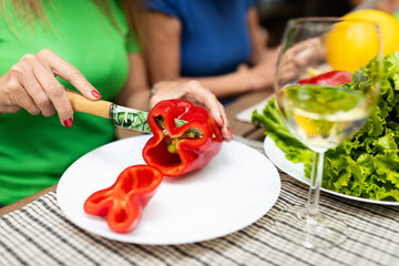 A person in a green shirt cuts a red bell pepper in a casual dining setting. Fresh lettuce is visible, alongside a glass of white wine, indicating a relaxed lunch atmosphere.