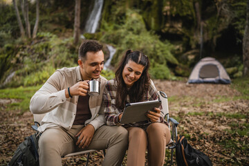 Couple sit in front tent in the forest and use digital tablet